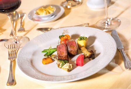 مطعم فندقي في GRAND HOTEL Zell am See طبق رئيسي مرتب بعناية على صحن أبيض على طاولة مُجهزة مع كأس نبيذ وأدوات مائدة.