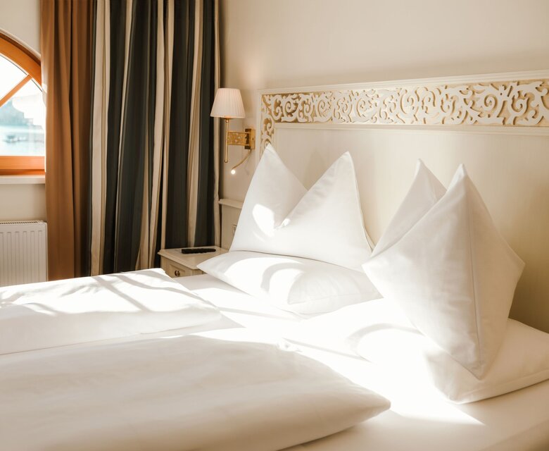 Ein helles Hotelzimmer mit gemachtem Bett, dekorativem Kopfteil und Blick durch das Fenster auf den See.