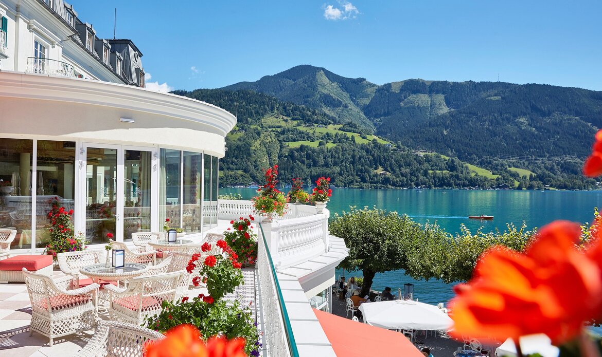 A Seebar terrace with white tables and chairs, flowers, and a view over Lake Zell and the mountains.
