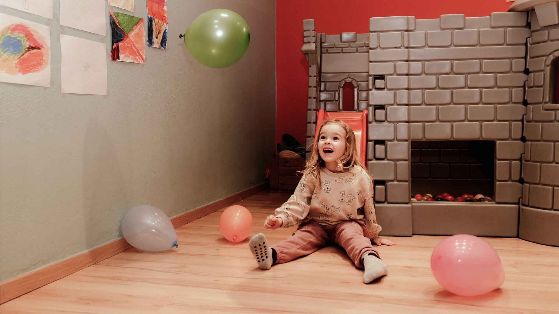 Ein kleines Mädchen sitzt zwischen Luftballons im Spielbereich mit Spielburg und Kinderzeichnungen.