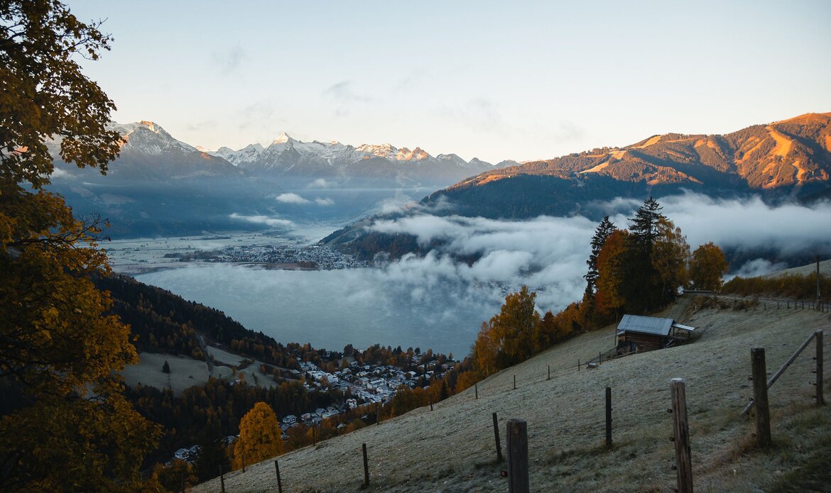Blick über den Zeller See mit Nebel im Tal, Bergen im Morgenlicht und einem Hang mit Zaun im Vordergrund.