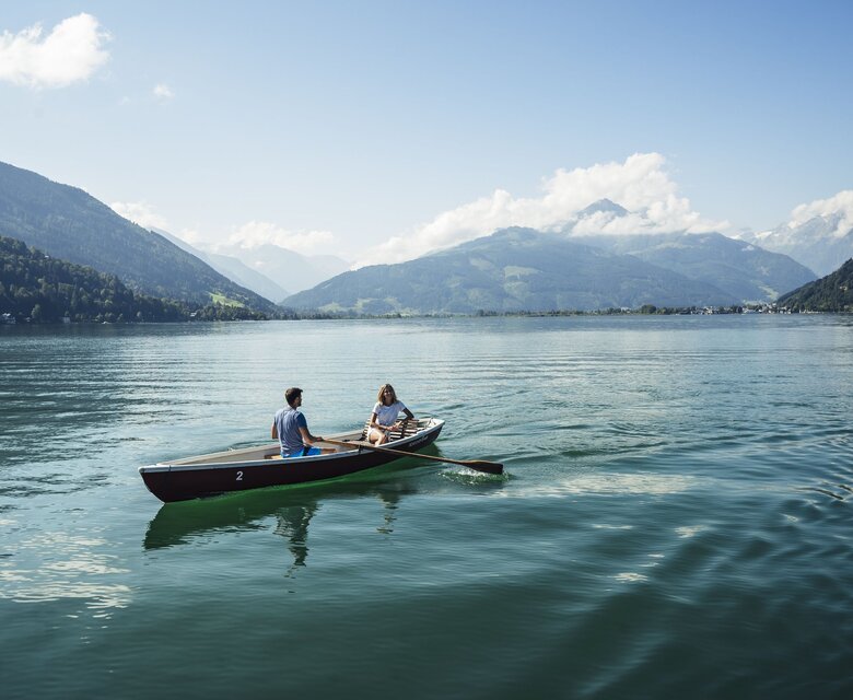 Zwei Personen sitzen in einem Ruderboot auf dem ruhigen Zeller See vor Bergkulisse im Sommer.