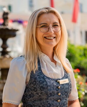 A smiling female staff member with a name badge stands outdoors in front of a blurred background.