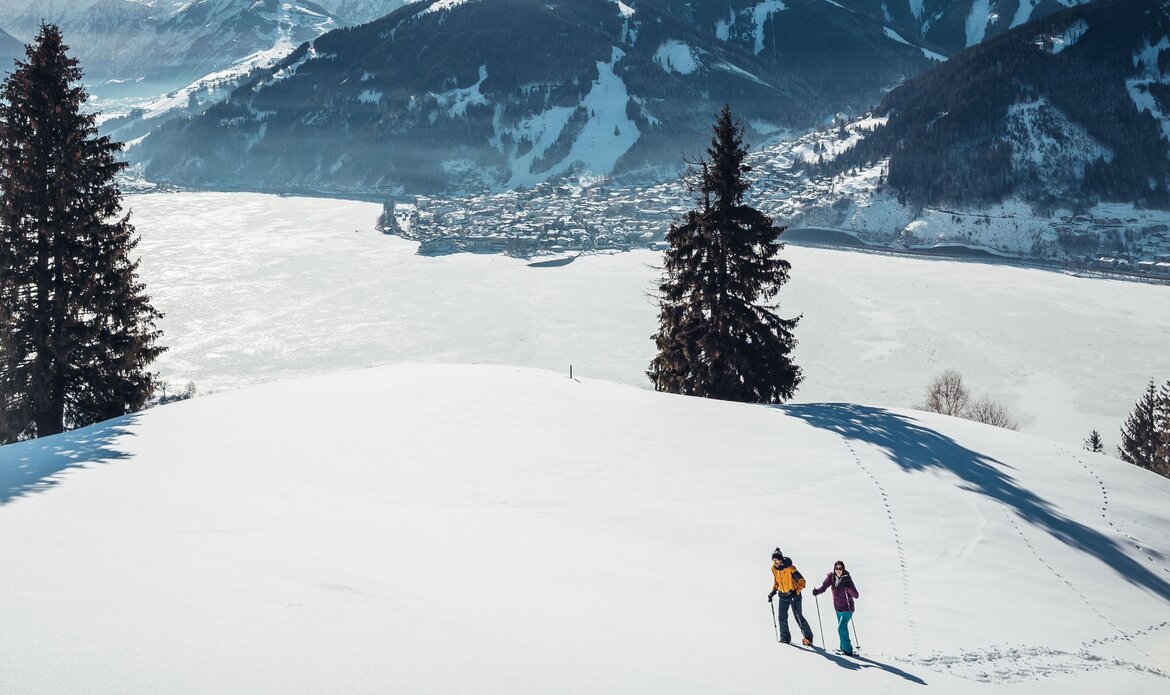 Two people walk across a snowy slope above the frozen lake with mountain views in the background.