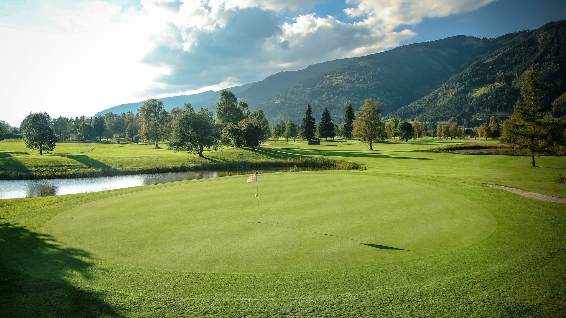 Ein gepflegtes Grün auf dem Golfplatz liegt vor einem Teich und einer Bergkulisse im Morgenlicht.