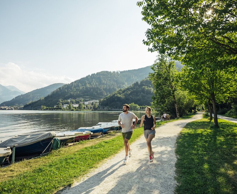 Zwei Personen joggen auf einem Uferweg am Zeller See, nebenan liegen Boote am ruhigen Wasser.