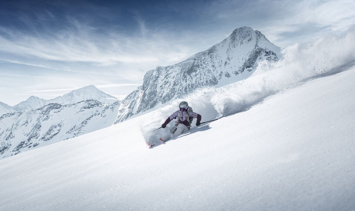 A skier carves through deep powder snow with a steep, snow-covered mountain behind them.
