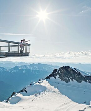 Zwei Personen stehen auf einer Aussichtsplattform über einer verschneiten Berglandschaft mit Blick auf die Alpen.