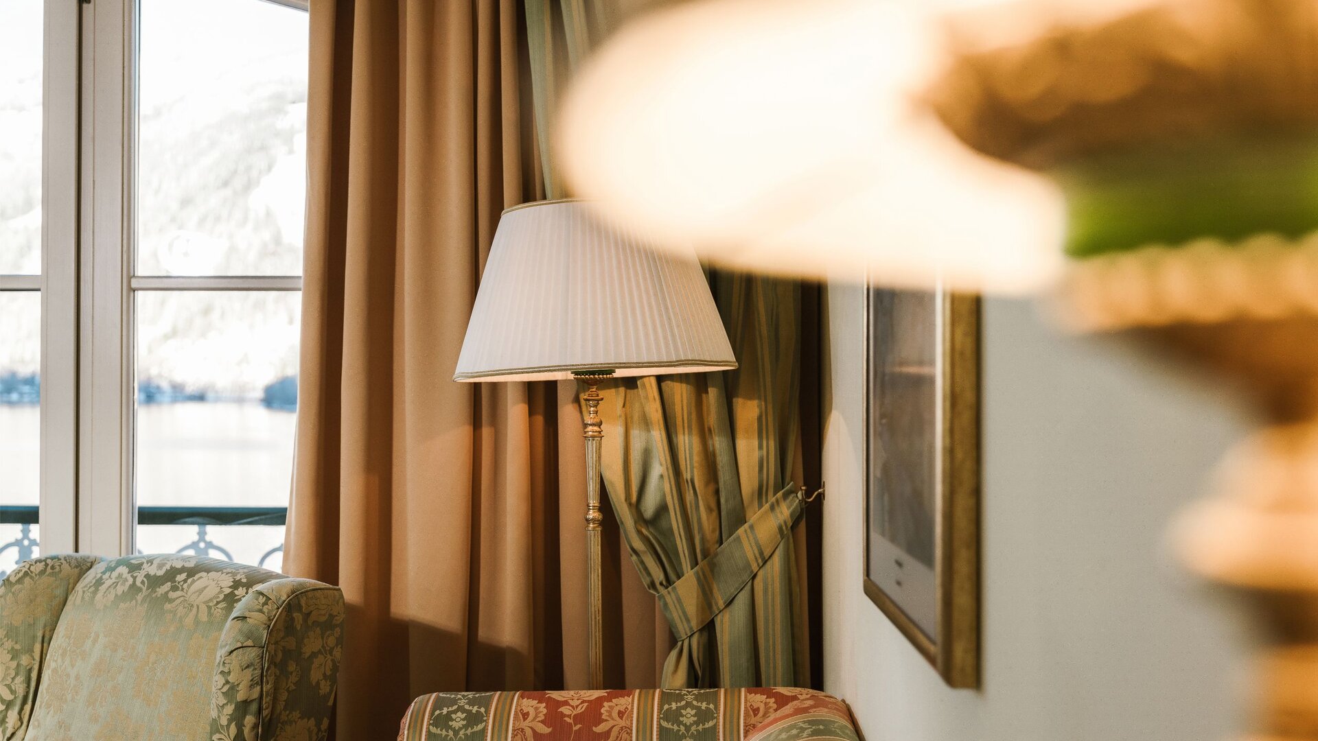 Ein Fenster mit Blick auf den See, Vorhänge, eine Stehlampe und ein gepolsterter Sessel im Hotelzimmer.