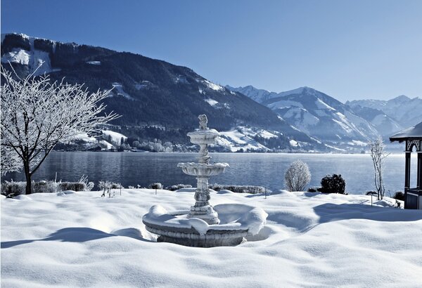 Ein verschneiter Garten mit steinerner Brunnenanlage liegt vor dem Zeller See und den Bergen.