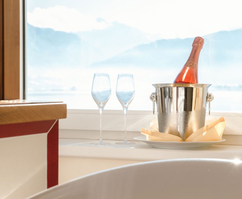 Champagner in einem Eiskübel mit zwei Gläsern am Fenster, im Vordergrund ein Wannenrand und der helle Seeblick.