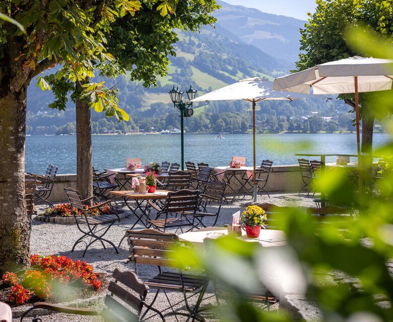 Auf der Restaurantterrasse stehen Tische, Stühle und Sonnenschirme mit Blick auf den See und die Berge.