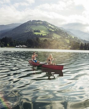 Zwei Personen paddeln in einem roten Kanu auf dem Zeller See vor grünen Bergen.