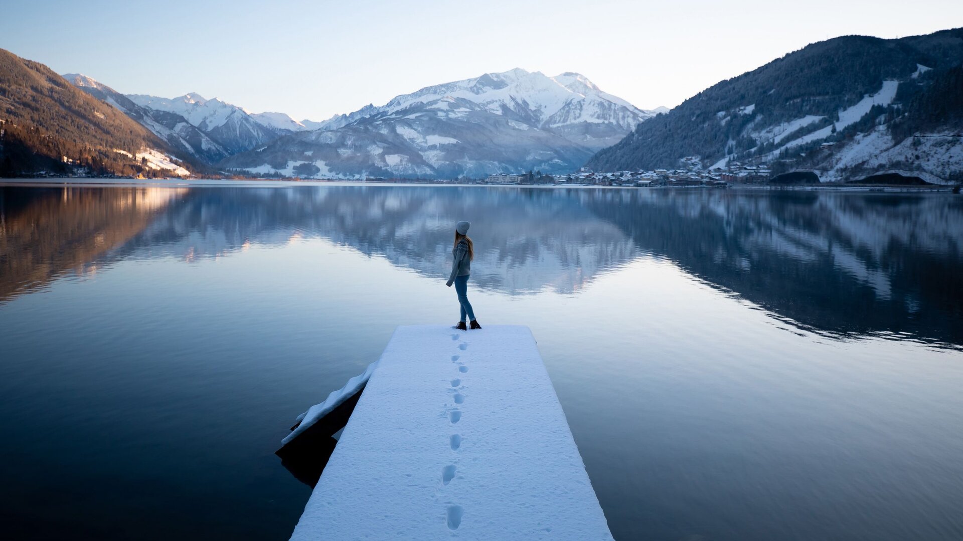 A person stands on a snow-covered jetty by the calm Lake Zell, with mountains reflected in the water behind them.