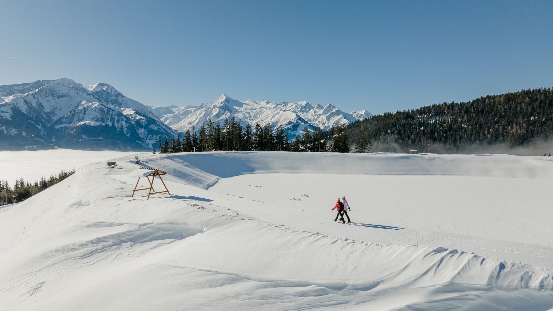 A person walks with poles across a snowy area with mountain views and a clear sky in the background.