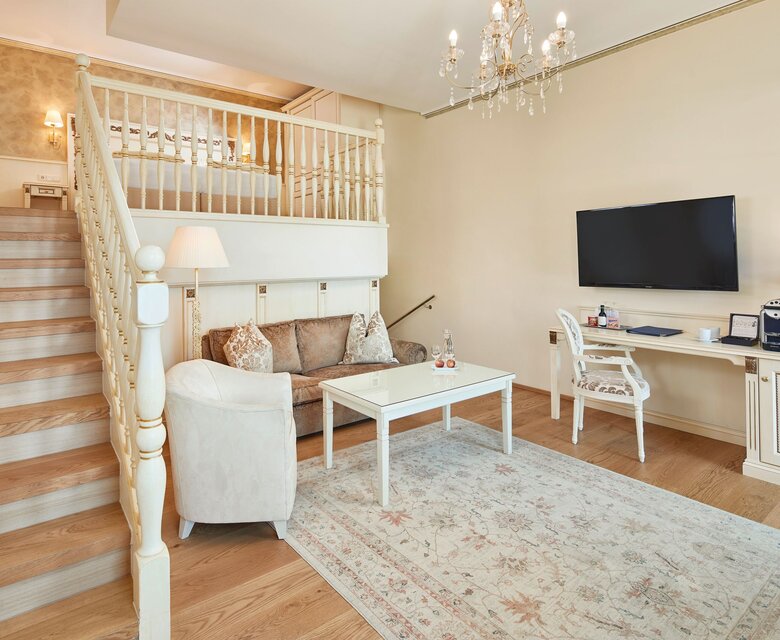 Ein heller Wohnbereich einer Suite mit Sofa, Schreibtisch, Treppe zur Galerie und einem Kronleuchter.