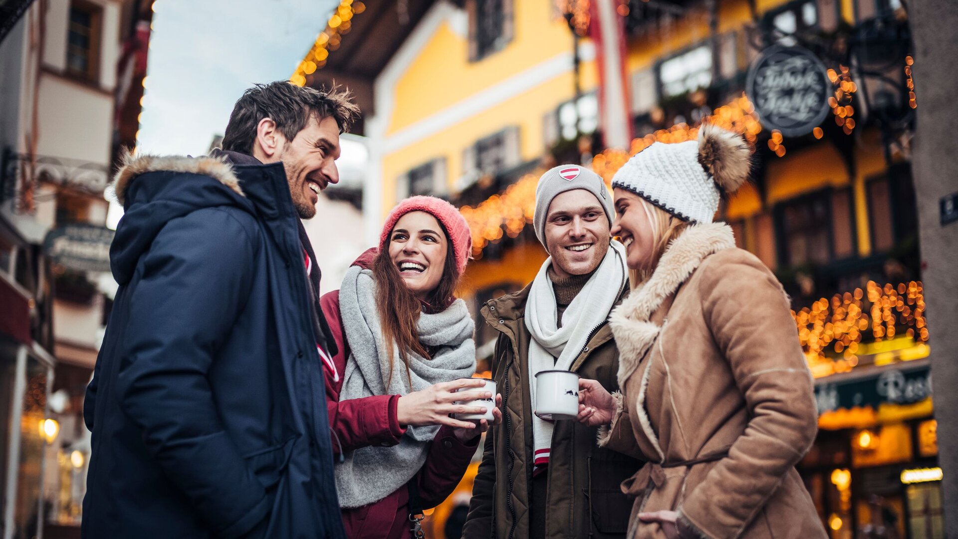 Four adults chat with hot drinks in front of festively lit houses in wintery Zell am See.