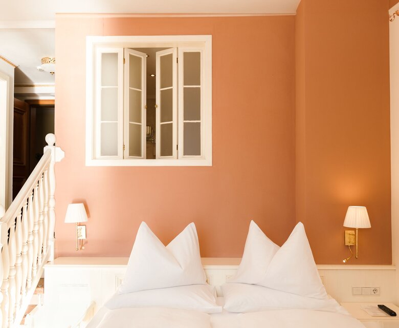 Ein helles Hotelzimmer mit Doppelbett, orangefarbener Wand, Treppe und Fenster im GRAND HOTEL Zell am See.