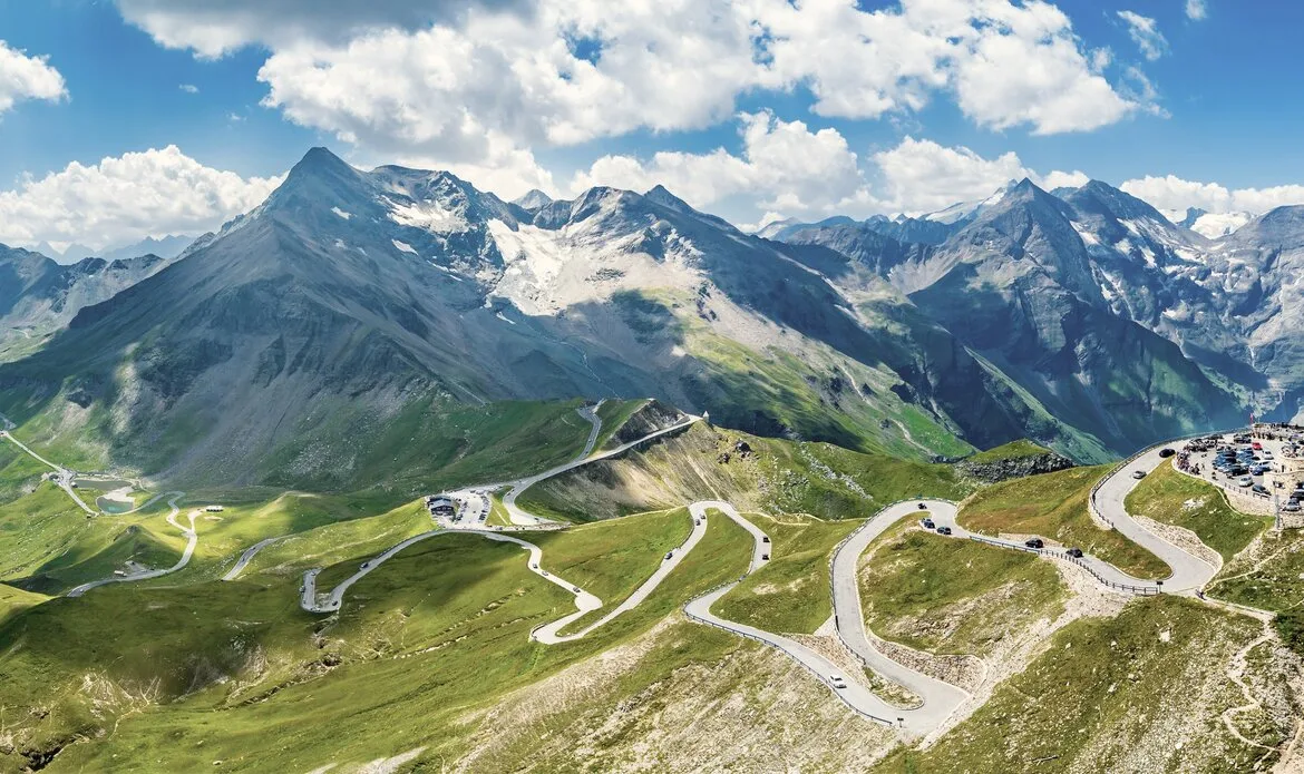Eine kurvige Panoramastraße führt über grüne Berghänge in einer alpinen Landschaft.