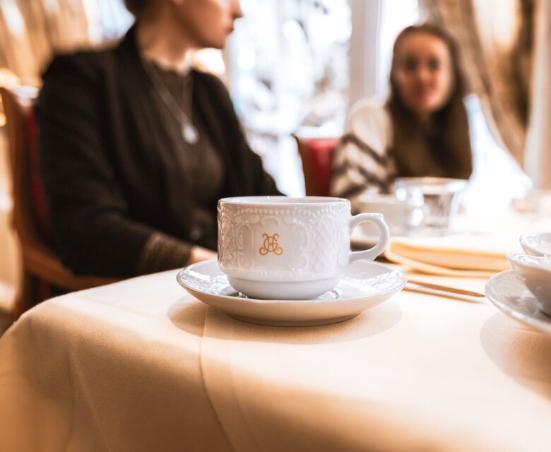 Eine Kaffeetasse mit Untertasse steht auf einem gedeckten Tisch im Hotelrestaurant; im Hintergrund sitzen Gäste.