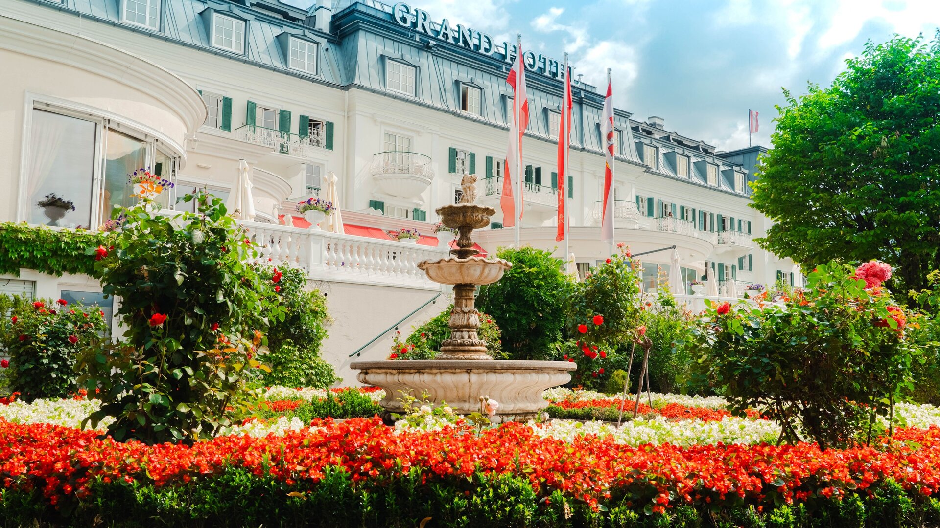 A fountain stands in a landscaped garden in front of the Grand Hotel, with flower beds and red flags nearby.