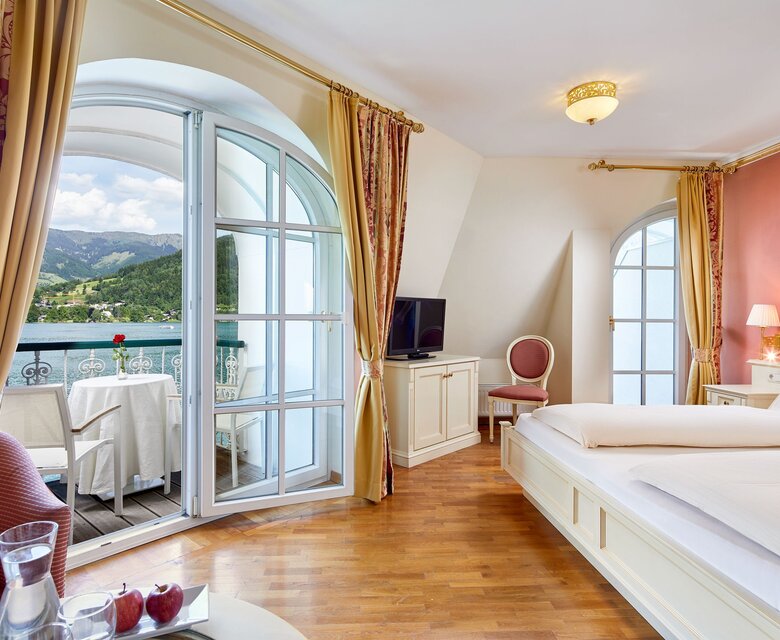 Ein helles Hotelzimmer mit großem Bett, Holzboden und offenem Balkon mit Blick auf den See.