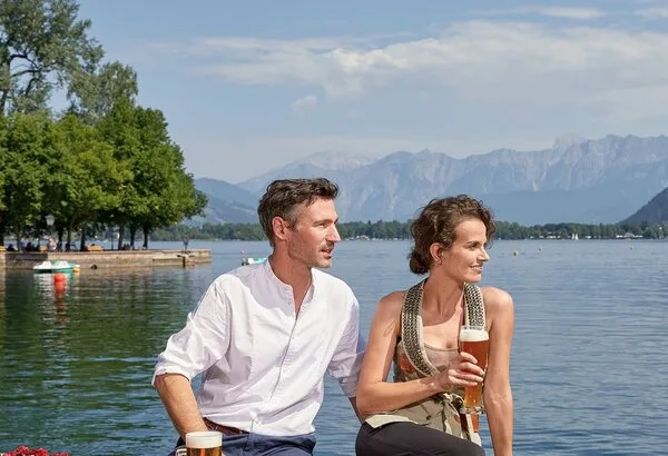 Ein Paar sitzt mit Bier und Speisen an einem Tisch direkt am Seeufer mit Bergblick.