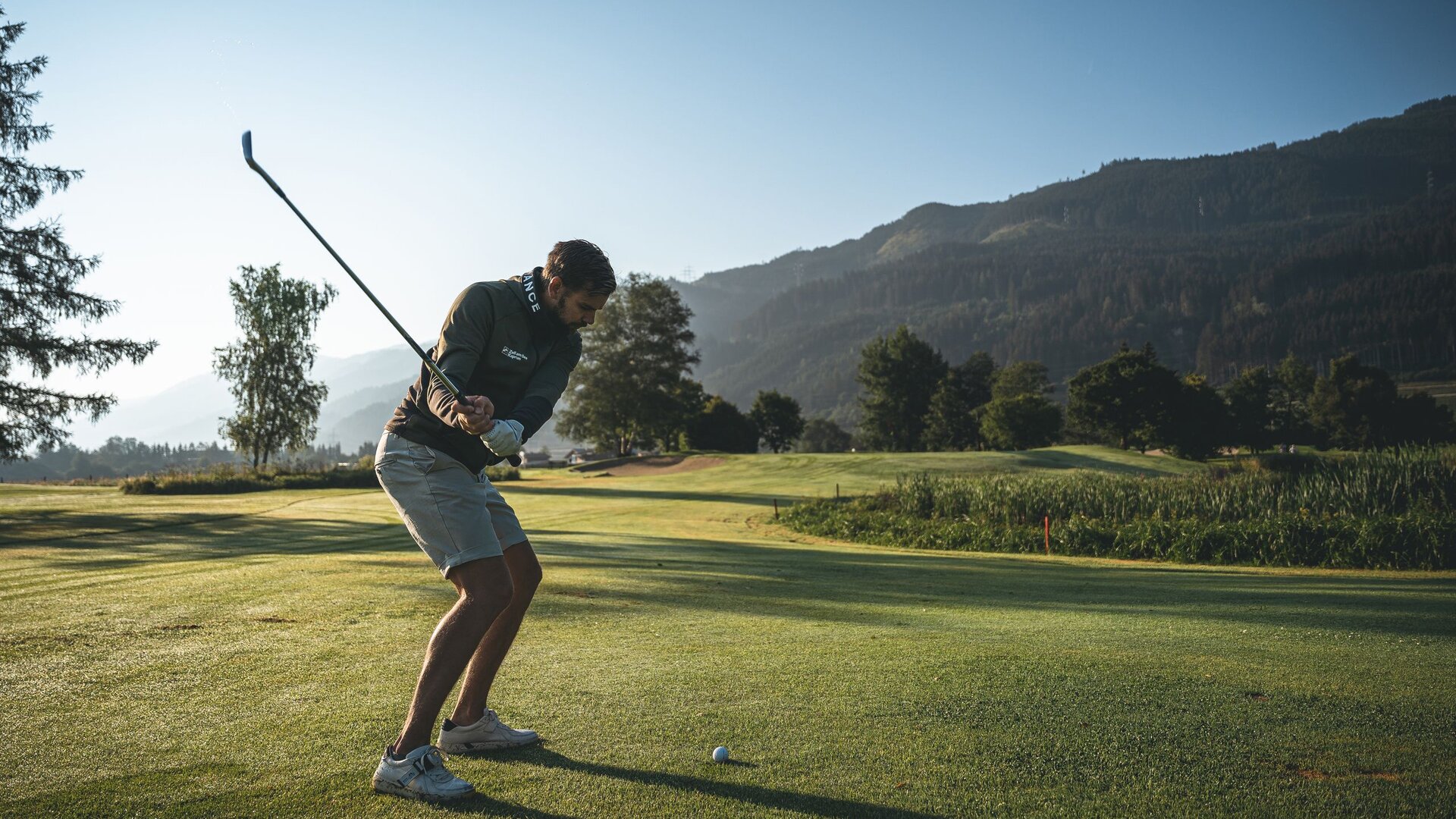 Ein Golfer bereitet auf dem Golfplatz seinen Abschlag vor, im Hintergrund liegen Berge und Morgenlicht.