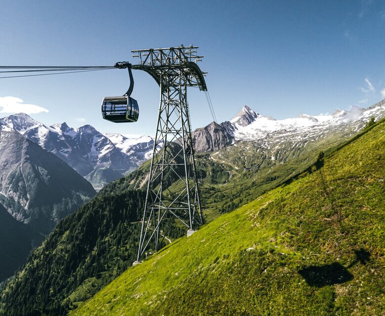 Eine Seilbahn fährt über eine grüne Bergflanke vor den schneebedeckten Gipfeln bei Zell am See.