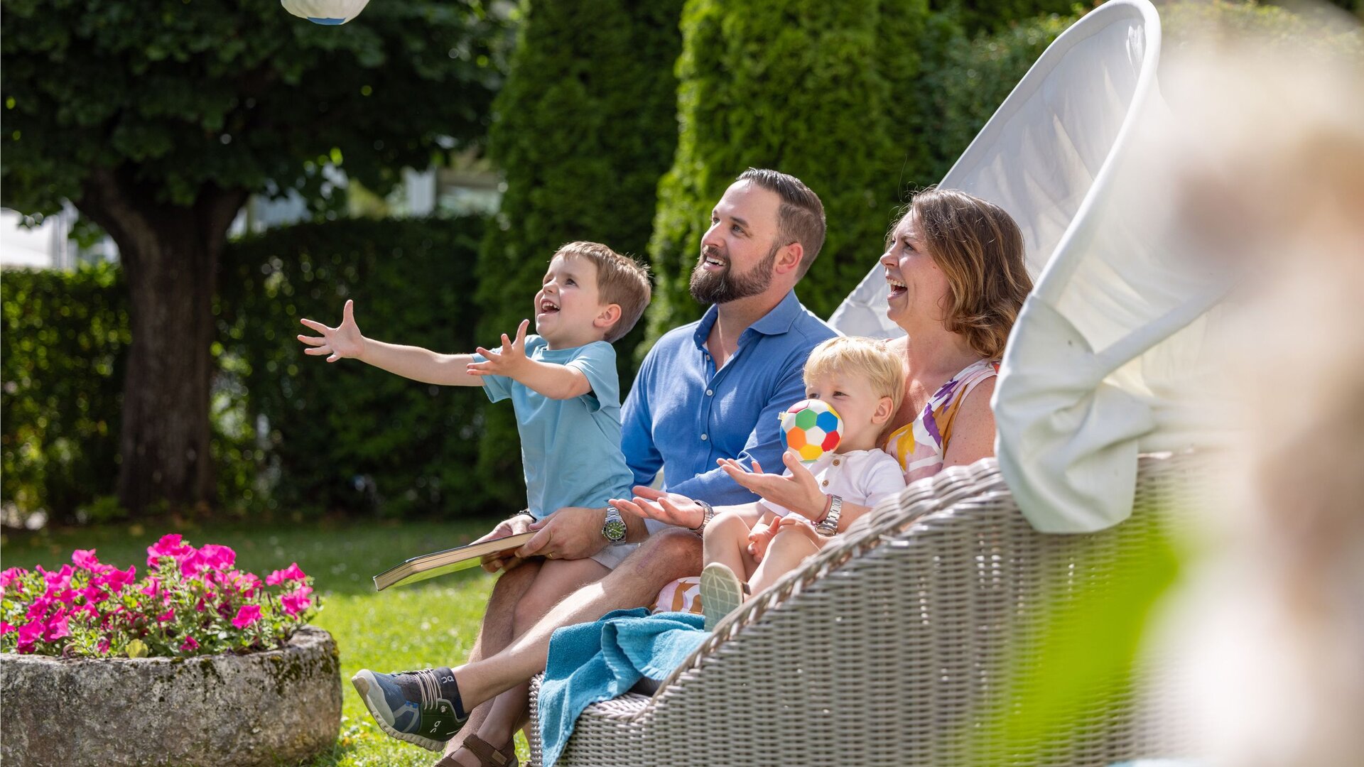 Eine Familie sitzt im Garten auf einer Lounge, während ein Junge nach oben greift und ein kleines Kind ein buntes Spielzeug hält.