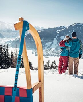 Ein Paar steht im Schnee und blickt über das winterliche Tal von Zell am See mit Bergen und See im Hintergrund.