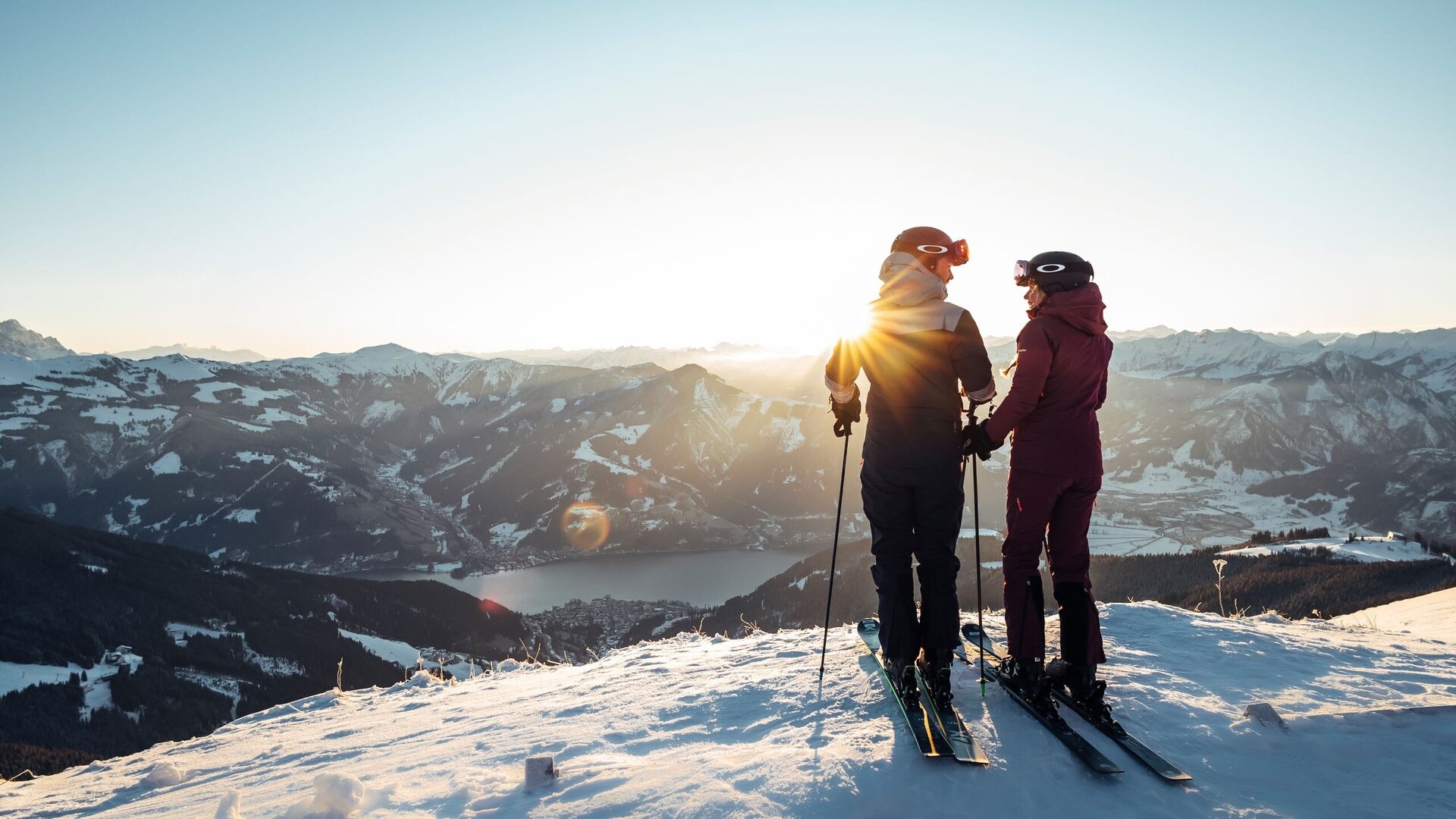 Two skiers stand on a snowy summit and look out over the mountain landscape near Zell am See.