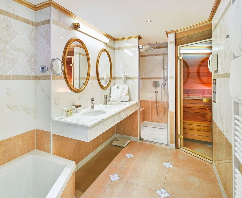 Ein helles Badezimmer mit Wanne, Doppelwaschtisch und Glasdusche; daneben führt eine Tür in eine private Sauna.