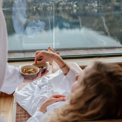 Eine Person im Bademantel frühstückt am Fenster mit Blick auf Wasser und Berge.