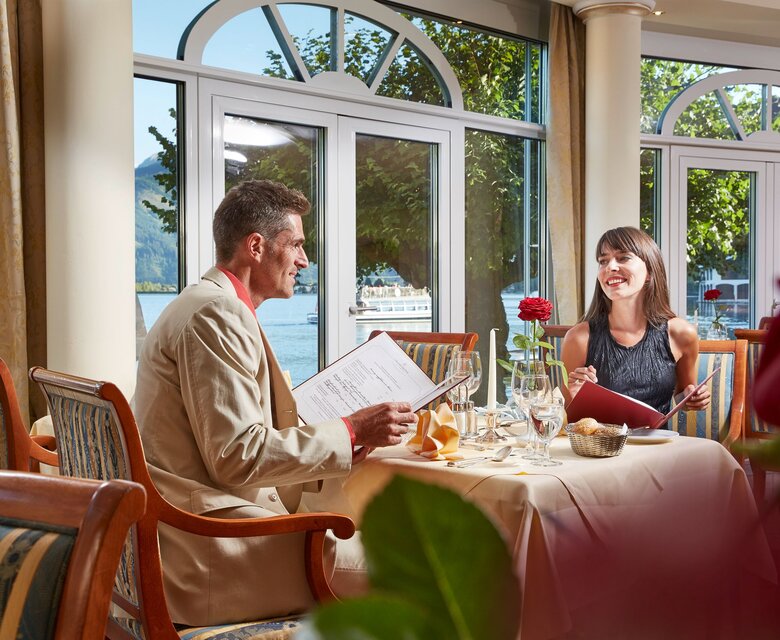 Ein Paar sitzt im Hotelrestaurant am Tisch mit Menükarten und Blick auf den See.