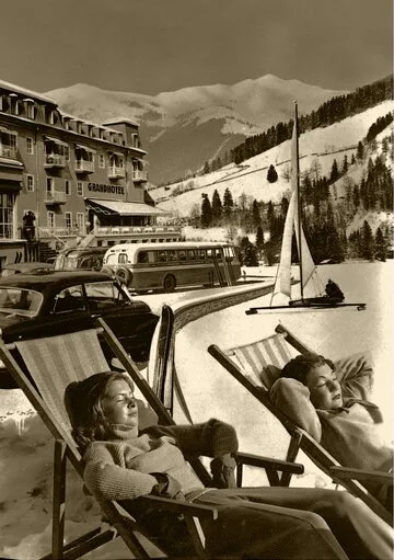 Zwei Personen liegen auf Liegestühlen vor dem GRAND HOTEL Zell am See, im Hintergrund Berge und ein verschneiter Hang.