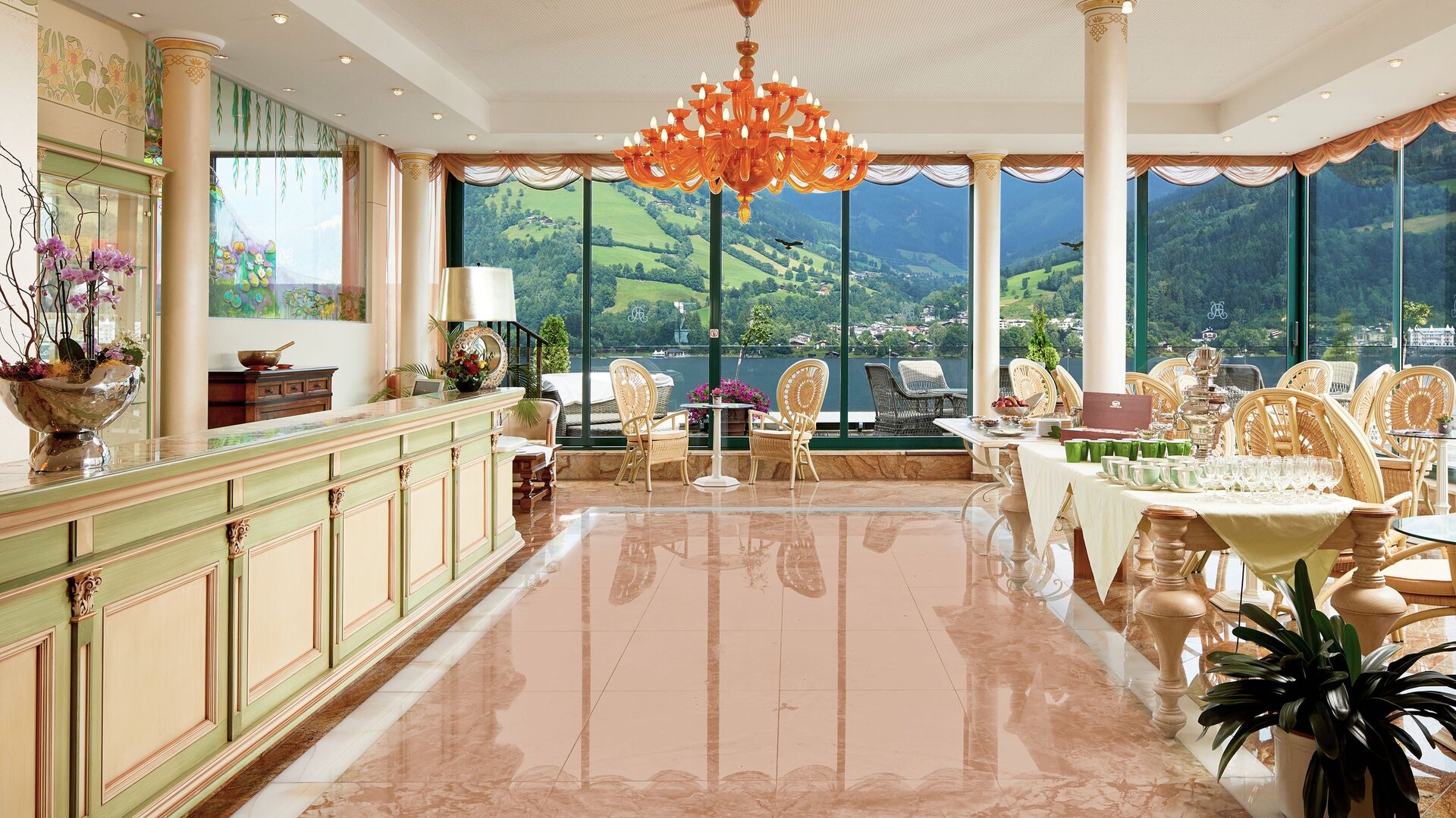 Ein heller Wellnessbereich mit Deckentischleuchten, Sitzgruppen und großen Fenstern mit Blick auf die Berge und den See.