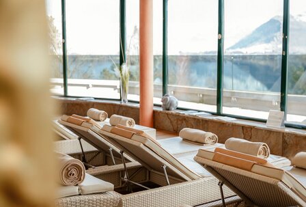 Spa-Liegen mit Seeblick im GRAND SPA Zell am See Mehrere gepolsterte Spa-Liegen stehen vor großen Fenstern mit Blick auf den See und die Berge.