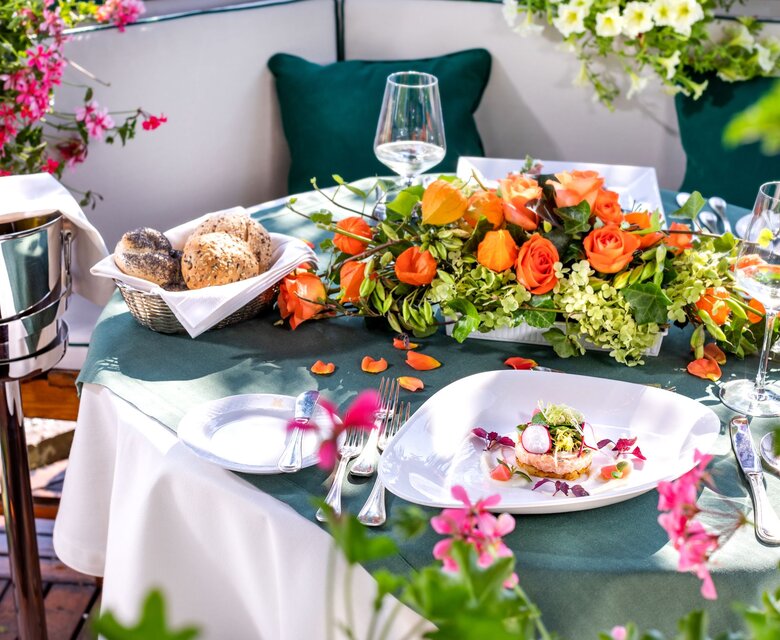 Ein gedeckter Tisch auf der Terrasse mit Blumenarrangement, Brotkorb, Weingläsern und einem angerichteten Teller.