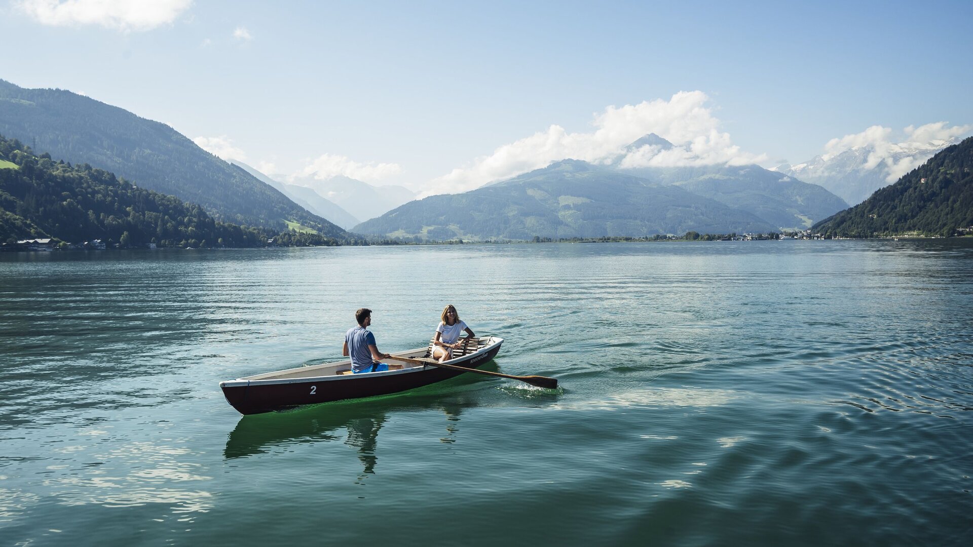 Zwei Menschen sitzen in einem Ruderboot auf dem Zeller See vor einer Bergkulisse.