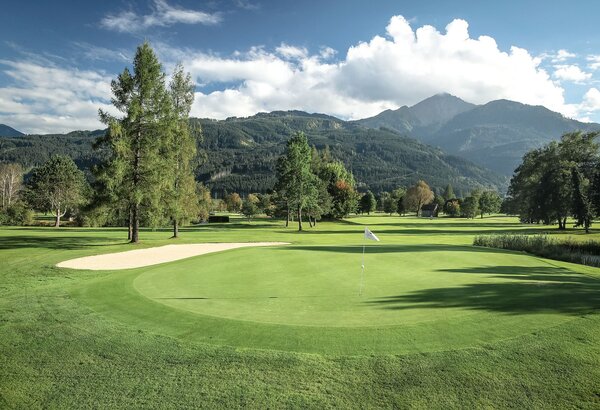 Ein gepflegtes Grün auf dem Golfplatz liegt vor einer Bergkulisse mit Bäumen und Himmel.