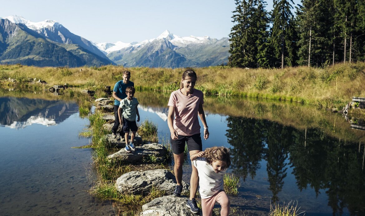 Eine Familie wandert auf einem schmalen Steg durch einen Bergsee mit Alpenkulisse.