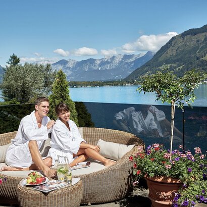 Ein Paar sitzt im Bademantel auf einer Lounge-Terrasse mit Blick auf den Zeller See und die Berge.