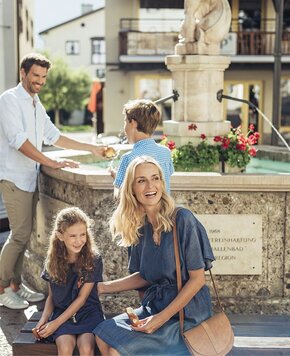 Eine Familie sitzt und steht an einem Brunnen im Sommer vor einem Gebäude in Zell am See.