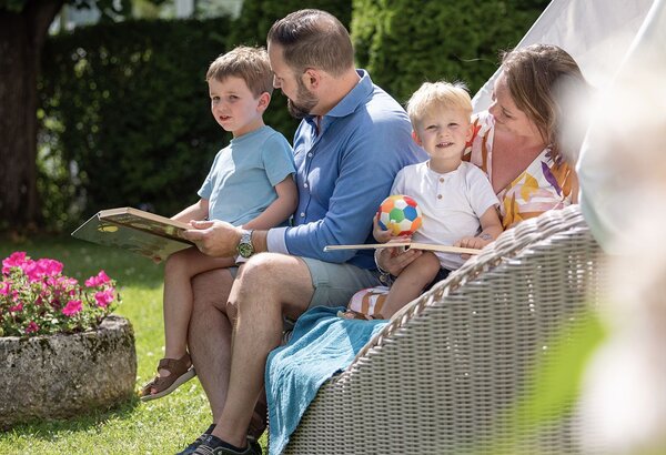 Eine Familie sitzt auf einer Gartenterrasse im Grünen, ein Mann liest den Kindern vor.