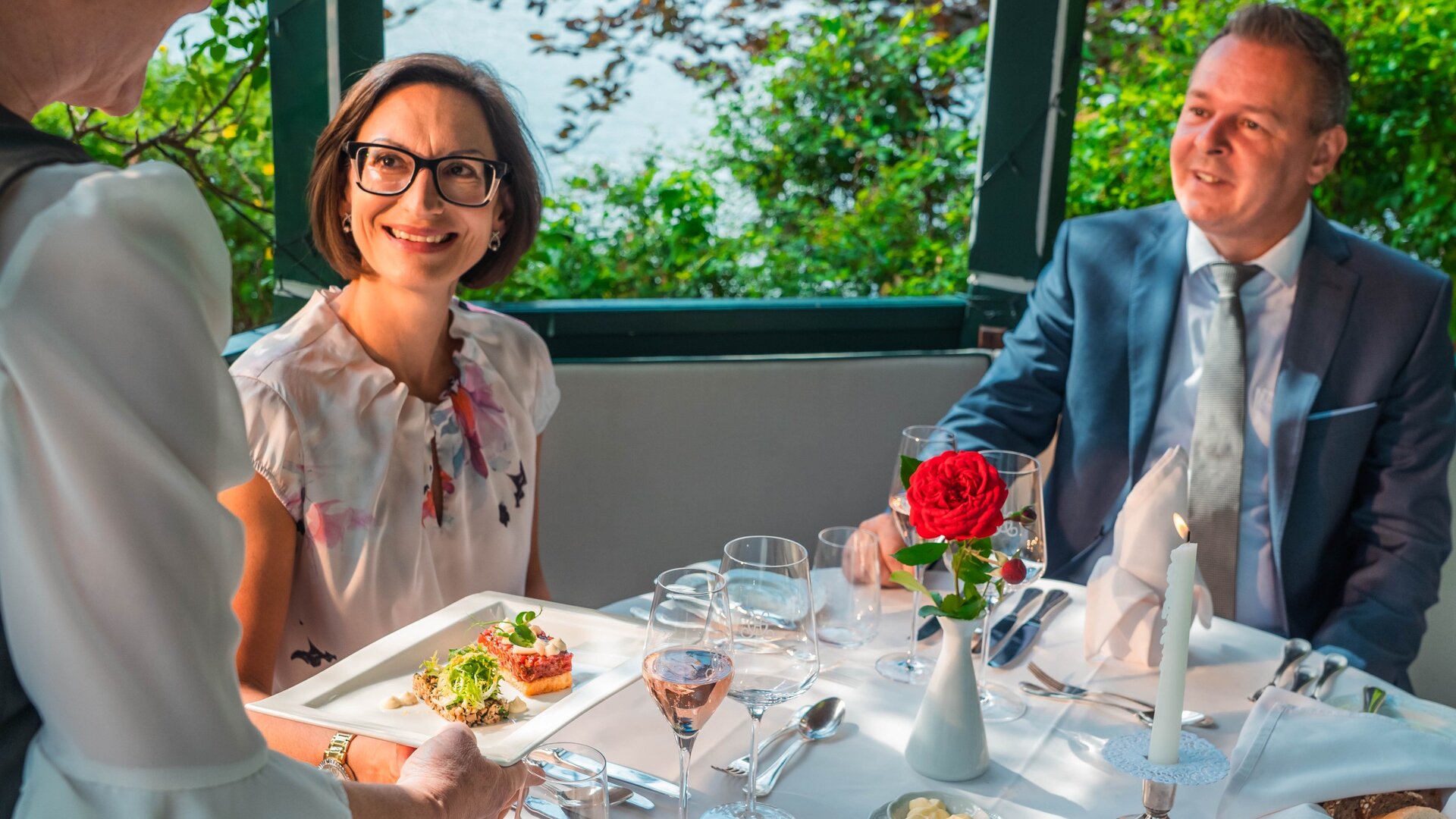 Zwei Gäste sitzen an einem gedeckten Tisch im Hotelrestaurant und sprechen mit einer Servierkraft.