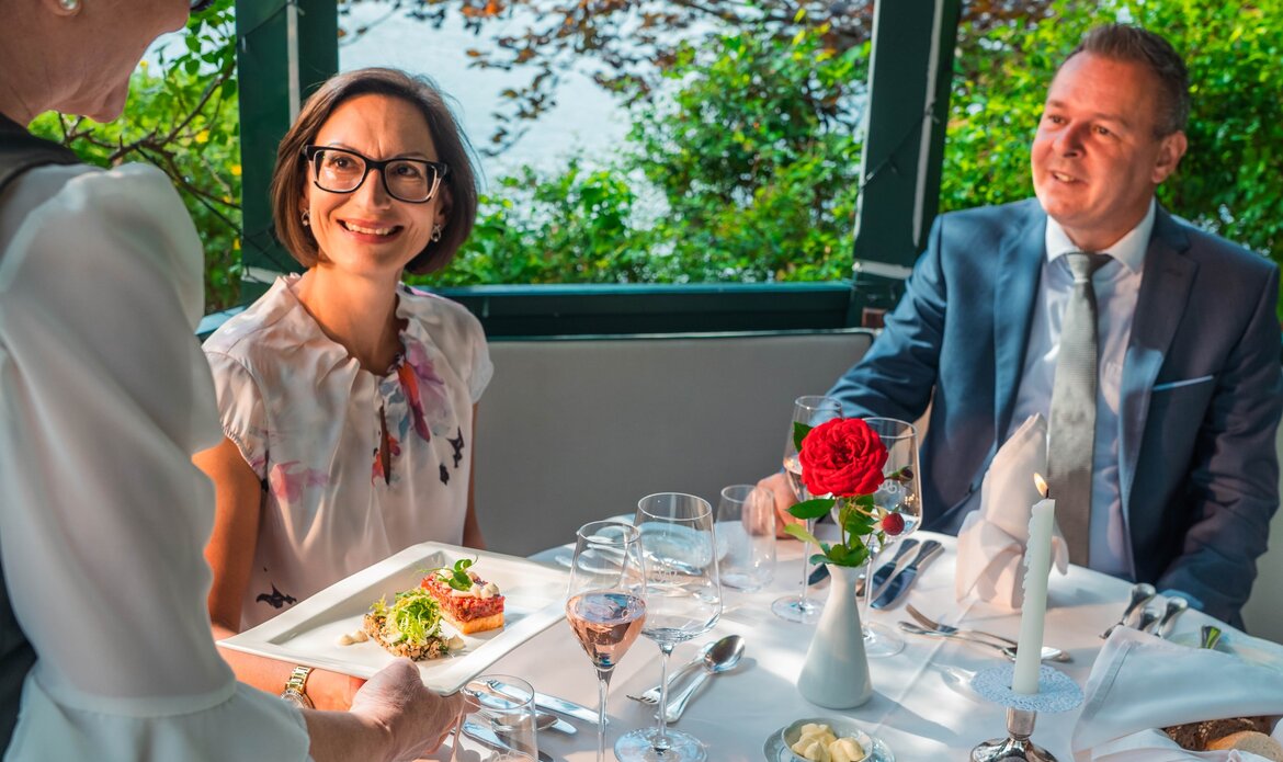 Zwei Gäste sitzen an einem gedeckten Tisch im Hotelrestaurant und sprechen mit einer Servierkraft.