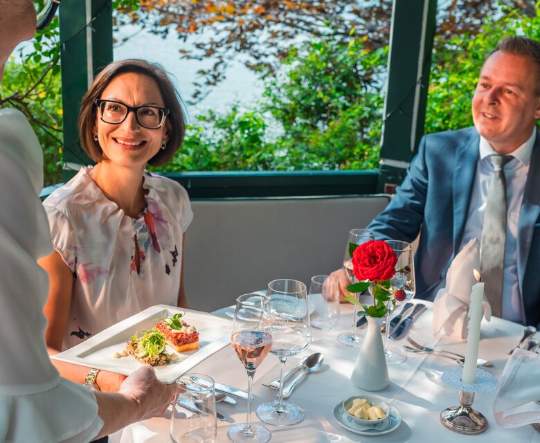 Zwei Gäste sitzen an einem gedeckten Tisch im Hotelrestaurant und sprechen mit einer Servierkraft.