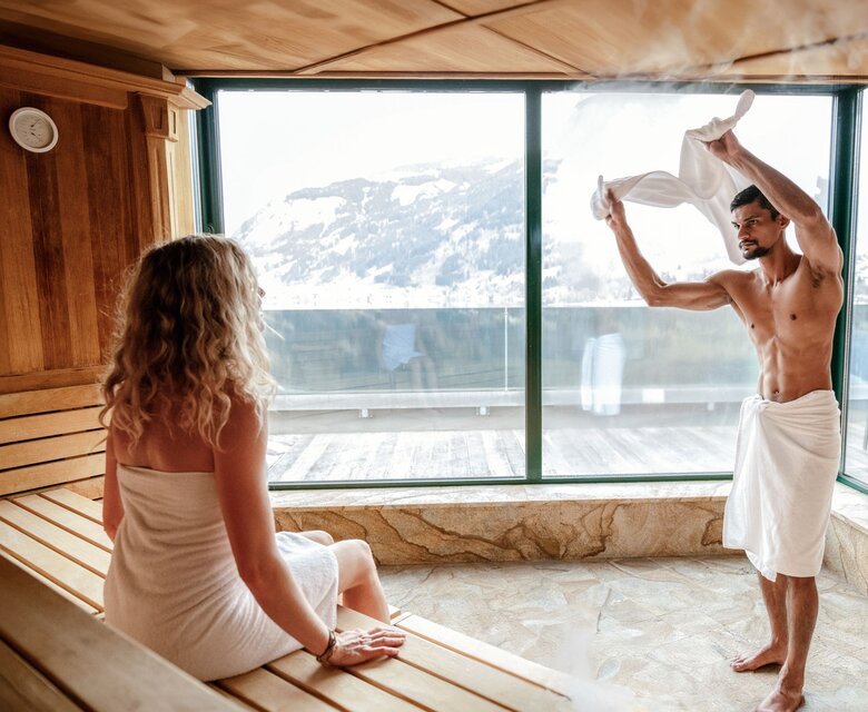 Ein Mann in der Sauna trocknet sich mit einem Handtuch ab, während eine Frau auf der Bank sitzt und zum See blickt.