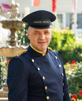 Ein Portier des GRAND HOTEL Zell am See steht im Freien vor einem Garten mit Springbrunnen.